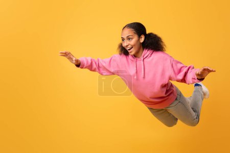 Téléchargez les photos : Adolescente noire excitée dans un sweat à capuche rose décontracté écartant les bras tout en volant, profitant de la liberté et du plaisir dans ses rêves sur fond de studio jaune, posant en plein air. Pleine longueur, espace libre - en image libre de droit