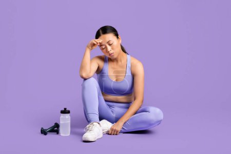 Téléchargez les photos : Épuisé asiatique femme athlète prendre pause, assis avec bouteille d'eau et haltère, essuyant la sueur de son front sur fond violet - en image libre de droit