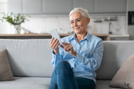 Photo for Senior Lady Wearing Eyeglasses Using Internet On Smartphone, Texting And Scrolling Social Media Application While Sitting On Couch In Modern Living Room At Home. Online Leisure, Communication - Royalty Free Image
