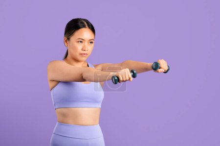 Téléchargez les photos : Focalisé jeune femme asiatique dans correspondant vêtements de sport étend les bras avec haltères pour la musculation sur fond violet - en image libre de droit