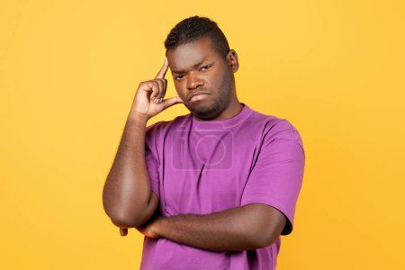 Téléchargez les photos : Homme noir mécontent réfléchi pensant à ses problèmes touchant la tête, regardant la caméra avec expression pénible, debout sur fond de studio jaune, portant un t-shirt violet - en image libre de droit