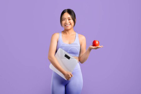 Foto de Mujer asiática feliz en ropa deportiva con confianza sosteniendo escamas en una mano y manzana roja fresca en la otra, simbolizando el enfoque equilibrado de la salud y la aptitud - Imagen libre de derechos