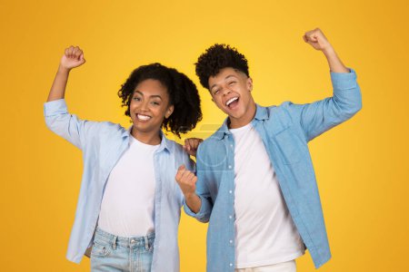 Photo for Two joyful black young people winners with arms raised in victory, sharing a triumphant moment, set against a vibrant yellow background, studio, exuding energy and enthusiasm - Royalty Free Image
