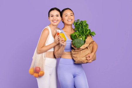 Foto de Dos mujeres alegres y diversas en equipo de entrenamiento sosteniendo bolsa de verduras frescas y jugo de frutas, simbolizando la dieta equilibrada y el deporte sobre fondo púrpura - Imagen libre de derechos