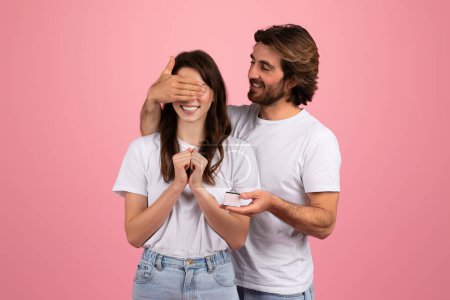 Photo for Happy young caucasian man covering woman eyes, hold gift box with red ribbon, isolated on pink background, studio. Surprise for anniversary, Valentine day, Birthday and proposal at romantic date - Royalty Free Image