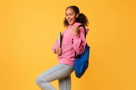 Téléchargez les photos : Heureuse jeune étudiante noire gestuelle oui tenant des matériaux de collège et portant un sac à dos, sur fond de studio jaune avec espace libre, célébrant la réussite scolaire à l'université - en image libre de droit