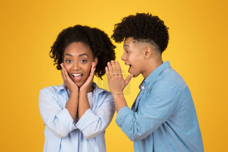 Téléchargez les photos : Une heureuse femme afro-américaine excitée avec les mains sur les joues dans la surprise tandis qu'un homme chuchote dans son oreille, les deux portant une tenue décontractée sur un fond jaune, studio - en image libre de droit