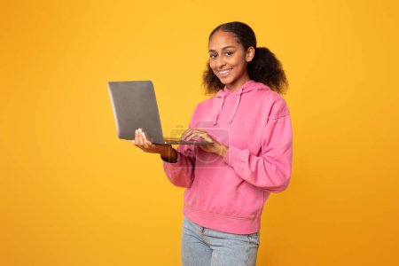 Téléchargez les photos : Joyeux jeune fille noire avec ordinateur portable navigation web pour les devoirs de l'école, engageant avec son carnet debout sur fond de studio jaune, souriant à la caméra. Surfer sur Internet étudiant sur ordinateur - en image libre de droit