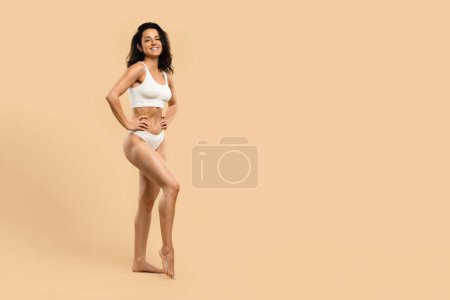 Photo for Beautiful young woman demonstrating her body sculpting achievements, attractive millennial female wearing white underwear set, embodying health and fitness, standing against beige studio background - Royalty Free Image