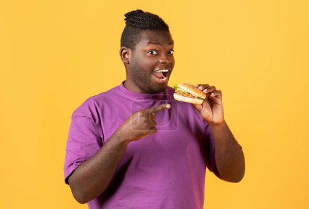 Photo for Portrait Of Funny Hungry Black Guy Pointing At Burger Eating Junk Food Posing Over Yellow Studio Background, Enjoying Unhealthy Nutrition And Cheat Meal. Binge Eating Habit - Royalty Free Image