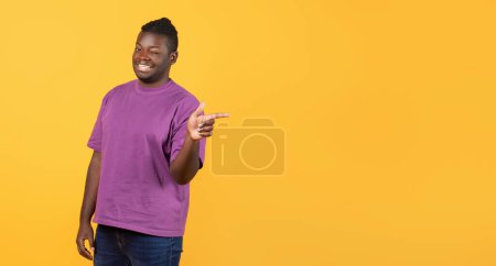 Téléchargez les photos : Joyeux jeune homme noir en t-shirt violet clignant des yeux et pointant du doigt, met en valeur l'espace libre pour l'offre de texte, debout sur fond de studio jaune. Panorama - en image libre de droit