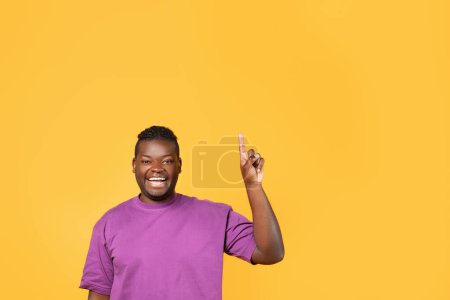 Téléchargez les photos : Jeune Afro-Américain excité avec un bras levé pointant vers le haut vers l'espace de copie, présentant un concept de grande idée, posant sur fond de studio jaune, signalant un moment d'inspiration - en image libre de droit