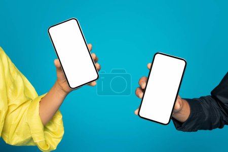 Téléchargez les photos : Deux mains tenant des smartphones à écran blanc tournés vers l'avant, l'une dans une manche jaune et l'autre dans une manche sombre, sur un fond turquoise contrasté, recadré, gros plan - en image libre de droit