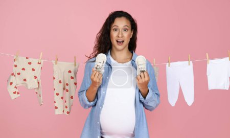 Photo for Overjoyed young pregnant woman in casual attire holds up small sneakers with baby clothes on a washing line in a pink backdrop, showing anticipation and happiness - Royalty Free Image