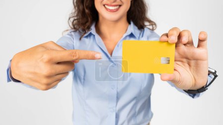 Foto de Primer plano de una mujer europea alegre en una blusa azul claro apuntando a una tarjeta de crédito amarilla que sostiene en su mano, con enfoque en la tarjeta y su expresión alegre, recortada - Imagen libre de derechos