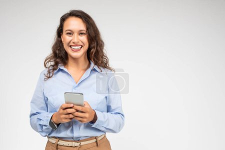 Photo for An engaging european young woman with curly hair and a bright smile looks up from her smartphone, wearing a blue shirt and khaki pants, suggesting connectivity and accessibility - Royalty Free Image