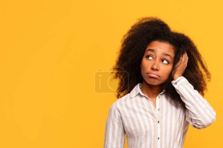 Téléchargez les photos : Contemplative jeune femme noire avec les cheveux bouclés regarde vers le haut, sa main dans ses cheveux, exprimant la réflexion sur fond jaune solide - en image libre de droit