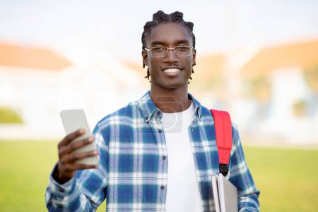 Foto de Sonriendo chico estudiante afroamericano se desplaza a través de los mensajes académicos en el dispositivo de teléfono inteligente, mirando a la cámara mientras posando con la mochila y gadget móvil universidad al aire libre. Tecnología y educación - Imagen libre de derechos