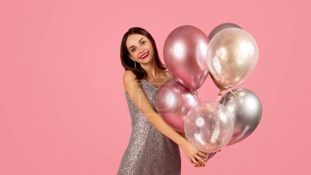 Téléchargez les photos : Femme caucasienne millénaire souriante dans une robe à paillettes glamour tenant un groupe de ballons métalliques et transparents, exsudant bonheur et célébration sur un fond rose - en image libre de droit
