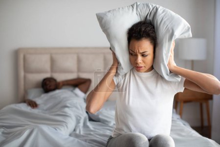 Photo for Loud Snoring Problem. Young frustrated black girlfriend covering ears with pillow in bed disturbed by her husbands loud noise, feeling upset at home bedroom. Empty space for text - Royalty Free Image