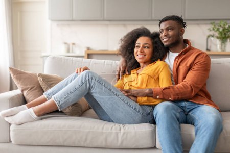 Téléchargez les photos : Joyeux mariage et bonne relation. Souriant afro-américains conjoints embrassant tout en se relaxant sur le canapé à l'intérieur, regardant loin bénéficiant du confort de leur salon intérieur moderne - en image libre de droit