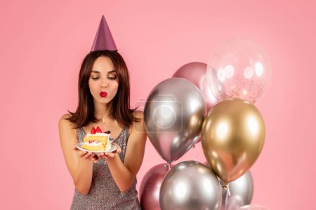 Photo for Playful happy young caucasian woman in a party hat blowing a kiss to a birthday cake, surrounded by metallic balloons, creating a festive atmosphere with her sequined dress - Royalty Free Image