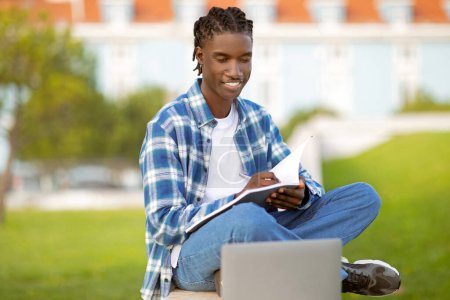 Photo for Cheerful young black student man studying with laptop and workbooks at park outside, writing notes engaging in e-learning and browsing on his computer. Digital education concept - Royalty Free Image