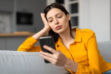 Photo for Worried young woman in yellow shirt, holding smartphone with pensive expression, sitting on gray couch in modern living room interior - Royalty Free Image