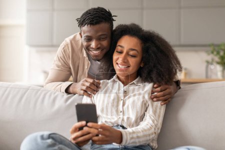 Téléchargez les photos : Couple noir joyeux partageant les médias sociaux sur le téléphone portable, heureuse famille afro-américaine assis sur le canapé, en utilisant un smartphone à la maison à l'intérieur, homme embrassant sa femme regardant l'écran de téléphone mobile - en image libre de droit