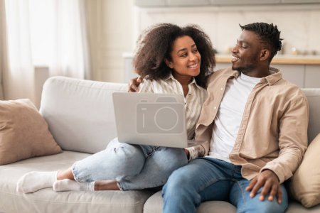 Téléchargez les photos : Les conjoints afro-américains milléniaux aiment regarder la vidéo en ligne sur leur ordinateur portable, se détendre sur le canapé dans le confort du salon à l'intérieur de la maison. Technologie en ligne, loisirs en informatique familiale - en image libre de droit