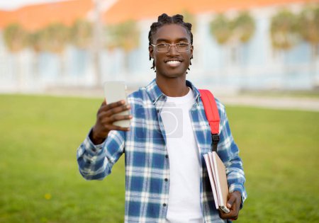 Photo for African American guy with backpack and workbooks uses cell phone outdoors, engaging with mobile gadget in the academic routine of his college, standing outside university campus - Royalty Free Image