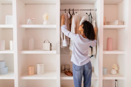 Photo for Curly-haired woman in casual attire thoughtfully arranging her stylish clothing in an open white closet in a room with soft pink walls - Royalty Free Image