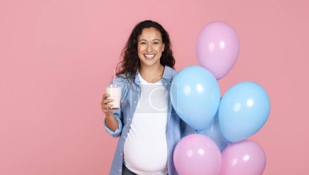 Téléchargez les photos : Jeune femme enceinte souriante avec un verre de lait tenant des ballons bleus et roses, buvant des produits laitiers pendant la fête de baby shower, isolée sur fond de studio rose, panorama avec espace de copie - en image libre de droit
