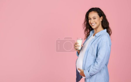 Foto de Saludable mujer embarazada bastante joven y feliz con ropa casual sosteniendo un vaso de leche, abrazando su gran barriga y sonriendo a la cámara, aislado en el fondo del estudio rosa, espacio de copia en blanco - Imagen libre de derechos