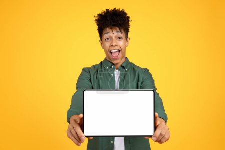 Foto de Joven afroamericano guapo alegre mostrando tableta digital con pantalla blanca en blanco y exclamando, recomendando oferta en línea, trato, fondo de estudio amarillo, maqueta, espacio de copia - Imagen libre de derechos