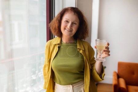 Photo for Senior woman enjoying glass of wine while standing by the window at home, smiling beautiful elderly lady posing in modern apartment setting, exuding elegance and relaxation, copy space - Royalty Free Image