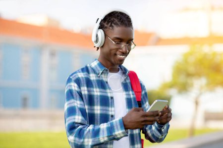 Téléchargez les photos : Étudiant afro-américain gars navigue application académique sur téléphone portable et écouter des livres audio avec écouteurs, profiter du mode de vie numérique tout en étudiant en plein air sur le campus - en image libre de droit