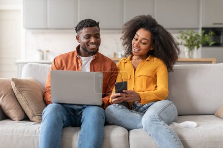 Photo for Great Online Offer. Happy Black Young Couple Reading Message On Smartphone And Using Laptop Computer, Sitting Together And Recommending Application Or Website On Sofa At Home - Royalty Free Image