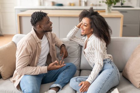 Téléchargez les photos : Heureux marié afro-américain couple parlant assis sur le canapé à la maison le week-end. Millennial conjoints noirs souriant à l'autre jouissant Flirt et conversation dans le salon confortable - en image libre de droit