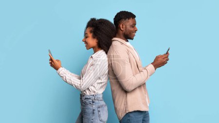Photo for Engrossed young black couple standing back-to-back, each absorbed in their own smartphone, highlighting the modern digital interaction on blue background - Royalty Free Image