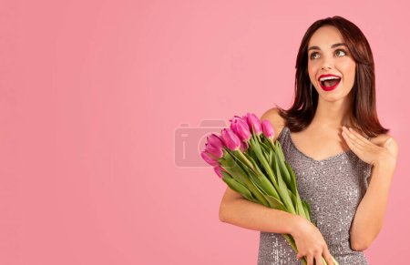 Photo for Surprised and delighted caucasian young woman in a glamorous sequined dress, holding a fresh bunch of pink tulips, looking away with a happy expression on a pink background - Royalty Free Image