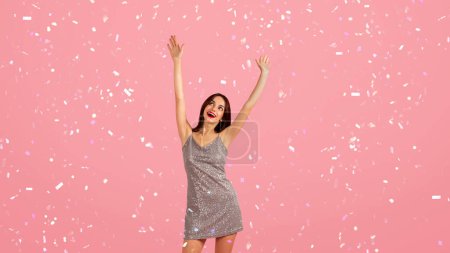Lebendige, fröhliche junge kaukasische Frau im Glitzerkleid mit breitem Lächeln, freudestrahlend erhobenen Armen, genießt Tanz, Spaß und Urlaub vor rosa Hintergrund mit Konfetti, Panorama