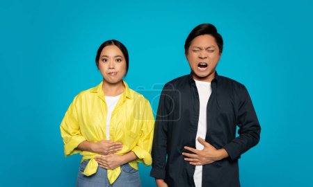 Photo for Concerned sad chinese young woman with hands clasped while a man beside her winces with a hand on his stomach, both against a turquoise background, studio, suggesting discomfort - Royalty Free Image
