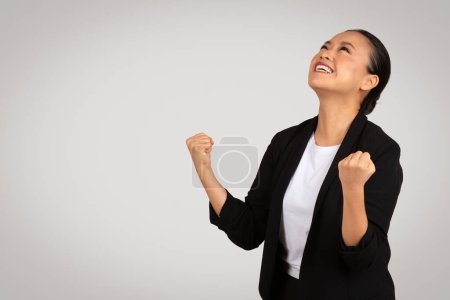 Téléchargez les photos : Éclatée heureuse femme d'affaires japonaise millénaire au sourire rayonnant lève les yeux, levant les poings dans un geste victorieux, symbolisant le succès sur un fond gris doux, studio - en image libre de droit
