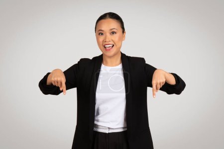 Téléchargez les photos : Enthousiaste heureuse femme d'affaires asiatique millénaire avec un large sourire pointant vers le bas avec les deux mains, soulignant quelque chose en bas, sur un fond gris neutre, studio - en image libre de droit