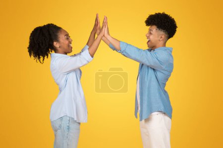 Téléchargez les photos : Jeune femme afro-américaine enthousiaste et homme haut-cinq avec des sourires brillants, célébrant le succès dans les vêtements décontractés sur un fond jaune optimiste - en image libre de droit