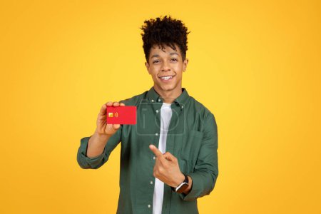 Photo for Cheerful stylish young black guy showing red plastic bank credit card and smiling, recommending contactless shopping, virtual banking, isolated on yellow studio background - Royalty Free Image