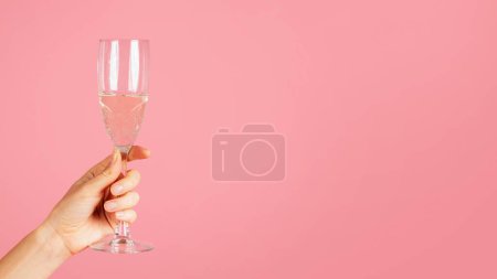 Téléchargez les photos : Une image en gros plan montrant une main tenant élégamment une flûte à champagne pétillante avec des bulles effervescentes, sur une toile de fond rose tendre avec amplement d'espace de copie, recadrée, panorama - en image libre de droit