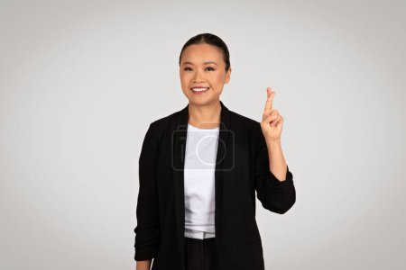 Téléchargez les photos : Une femme d'affaires asiatique souriante et souriante en veste noire avec une expression joyeuse, placée sur fond gris, pointe du doigt vers l'espace libre. Entreprise et recommandation de travail - en image libre de droit
