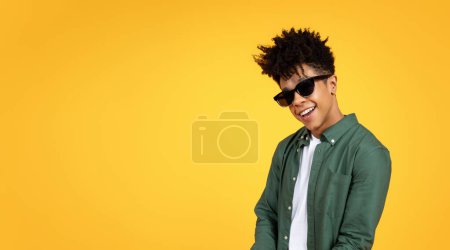 Foto de Elegante chico afroamericano joven con buen peinado con gafas de sol posando aislado en el fondo del estudio amarillo con espacio de copia en blanco para publicidad, web-banner - Imagen libre de derechos